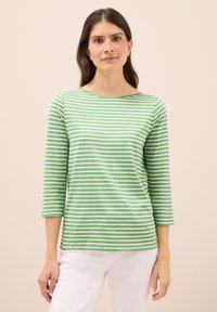 Zielona i biała w paski bluzka z długim rękawem o łódziastym dekolcie i dopasowanych rękawach, wykonana z miękkiego materiału. Noszona z białymi spodniami.