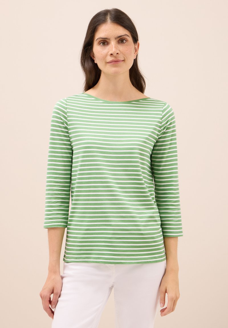 Zielona i biała w paski bluzka z długim rękawem o łódziastym dekolcie i dopasowanych rękawach, wykonana z miękkiego materiału. Noszona z białymi spodniami.