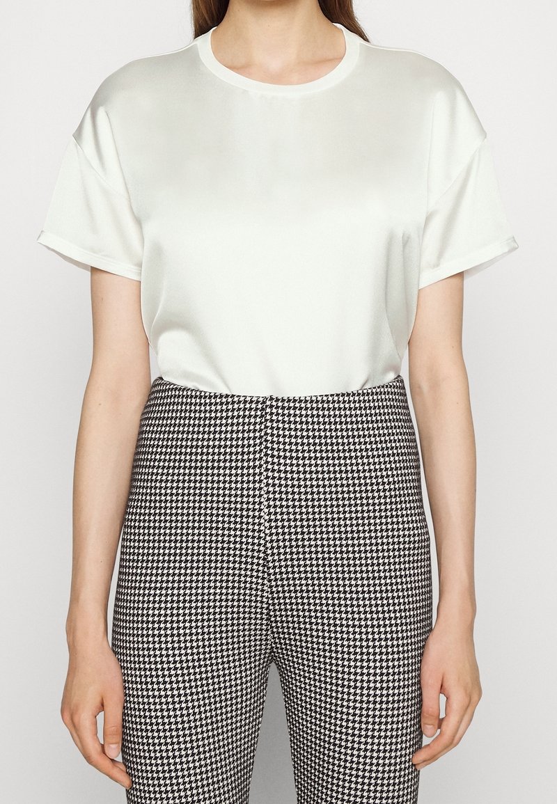 Femme portant un chemisier en satin blanc à manches courtes rentré dans un pantalon taille haute noir et blanc à motif pied-de-poule, sur un fond clair.