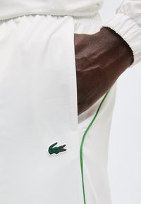 Σκούρο δέρμα χεριού στην τσέπη λευκού παντελονιού με πράσινη επένδυση και μικρό πράσινο λογότυπο κροκοδείλου κοντά στην τσέπη.