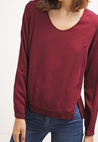 Burgunderfarbener Langarm-Top mit Rundhalsausschnitt. Weicher, glatter Stoff mit seitlichen Schlitzen am Saum. Kombiniert mit enganliegenden blauen Jeans.