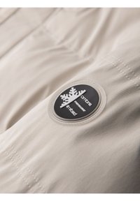 Gros plan sur un tissu beige avec un patch logo rond noir et blanc présentant un motif de flocon de neige et un texte cousu sur un vêtement.