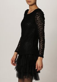 Blusa de encaje negra con mangas largas y escote redondo, combinada con una falda de tul negra con volantes. Detalle de encaje texturizado en toda la prenda.