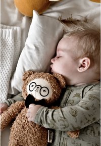 Soft teddy bear with glasses and a black bow tie held by a child in a light green patterned onesie, resting on white bedding.