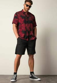 Zwart t-shirt met korte mouwen en een rood palmbladerenpatroon, gecombineerd met zwarte shorts en zwarte hoge sneakers, staand met handen in de zakken.