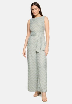 Nő világoszöld, ujjatlan, cikkcakk mintás overált visel, amely bő szárú, deréknál övvel megkötve, fehér háttér előtt áll.