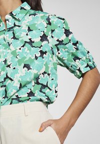 Chemise à manches courtes avec un motif floral vert, noir et blanc ; tissu léger, coupe décontractée, avec des manches froncées et une poche.