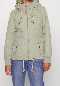 Ljusgrön, zip-up jacka med huva, dragsnören, två framfickor och ribbade ärmslut. Tillverkad av slätt, vattenavvisande material.