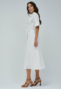 Abito midi bianco con maniche corte, colletto e dettaglio vita legato. Indossato con sandali con tacchi a strappi marroni. Tessuto liscio, design minimalista.