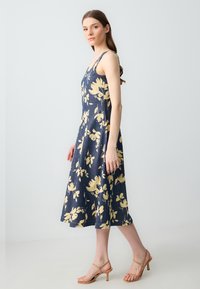 Blumenkleid aus navyfarbenem Stoff mit gelben Blüten, A-Linien-Silhouette, ärmelloses Design, knielange Länge, kombiniert mit tanfarbigen Riemchen-Highheels.