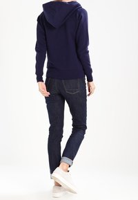 Sudadera azul marino con capucha ajustable, puños y dobladillo acanalados. Combinada con jeans de mezclilla oscuros y zapatillas blancas. Diseño minimalista.