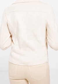 Dopasowana kurtka z jasno-beżowej sztucznej skóry z wysokim kołnierzem, dopasowanym krojem i delikatnym detalem szwów. Charakteryzuje się gładką powierzchnią i brakiem widocznych elementów metalowych.