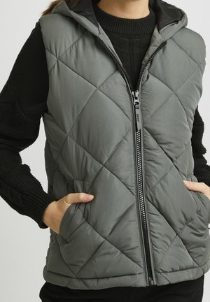 Person trägt eine graue gesteppte ärmellose Weste mit Frontreißverschluss und Taschen über einem schwarzen Strickpullover, Hände in den Westentaschen.