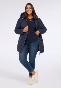 Abrigo puff en color azul marino con capucha, cierre de cremallera y bolsillos laterales. Acompañado de un suéter azul marino y jeans azules con acentos de tachuelas en el dobladillo.