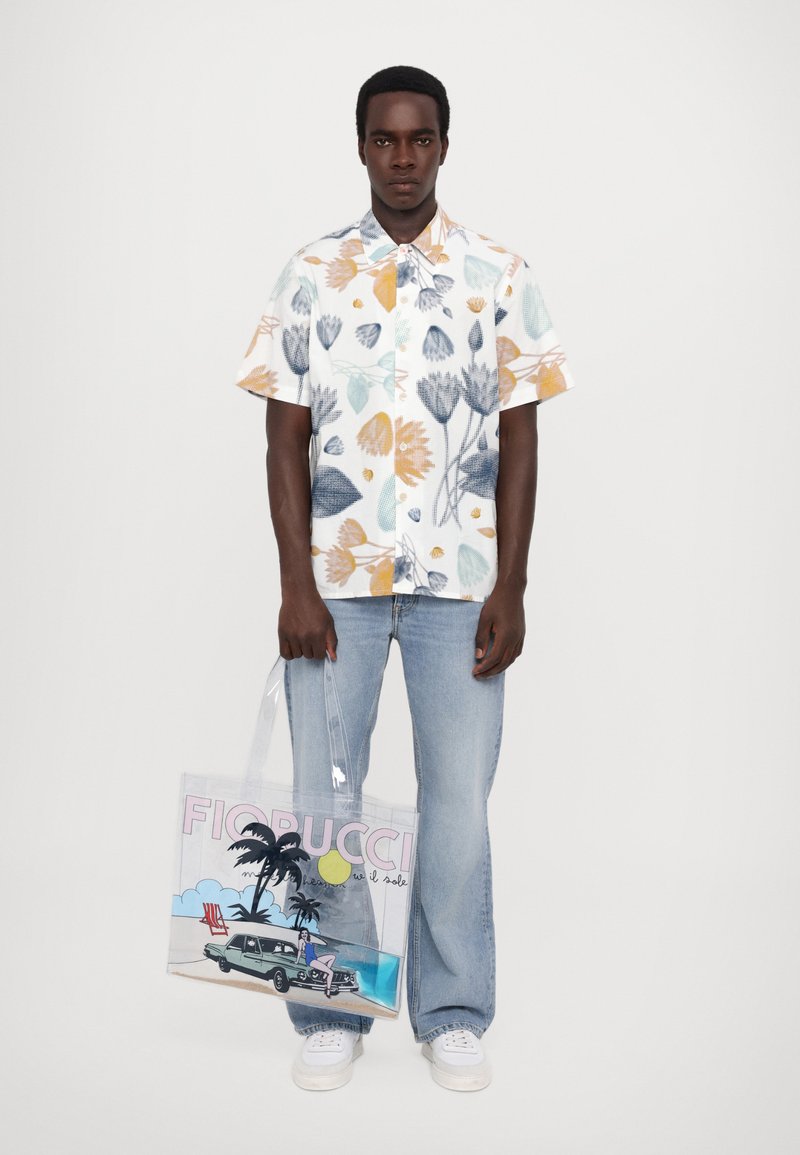 Chemise à manches courtes florale blanche avec des motifs bleus, oranges et verts ; associée à un jean léger bleu et un sac fourre-tout transparent avec un graphique.
