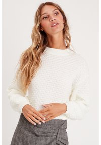 Pull en tricot blanc avec un motif texturé, col rond côtelé et manches longues, associé à une jupe à carreaux.