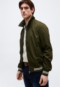 Jeune homme portant une veste zippée vert olive avec des poignets à rayures blanches, un pull léger, un jean foncé et une ceinture marron, debout devant un fond blanc.