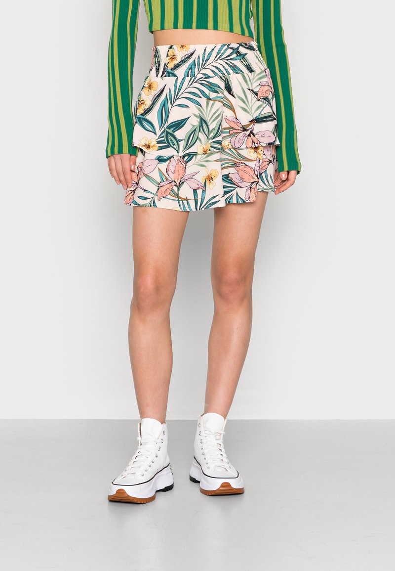 Saia mini floral com camadas de volantes, apresentando lírios cor-de-rosa e folhas verdes sobre um fundo creme. Combinada com botas brancas até ao tornozelo.