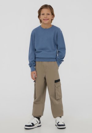 Felpa blu con scollatura rotonda e polsini a coste, abbinata a pantaloni cargo beige con tasche laterali e inserti neri con zip. Sneakers bianche.