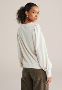 Ženska z kodrastimi lasmi, spetimi v nizek čop, z zlatimi obročastimi uhani, kremasto bluzo z dolgimi rokavi z teksturo in rjavimi žametnimi hlačami stoji pokončno iz strani.