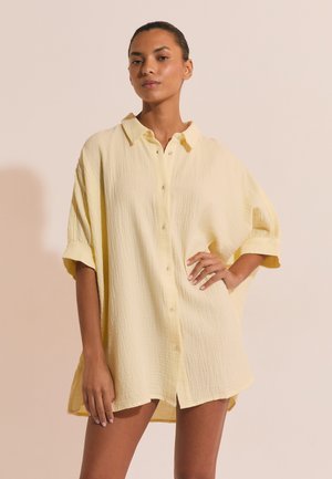 Kobieta z krótkimi włosami w luźnej, jasnżółtej sukience koszulowej z guzikami i podwiniętymi rękawami na beżowym tle.