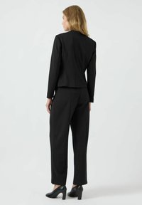 Černý oblek na míru s propracovaným sakem, rovnými kalhotami a jemným švem. Modelka má na sobě černé lodičky na vysokém podpatku.