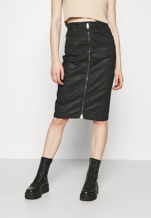 Woman wearing a black knee-length skirt with front zipper, beige cropped top, and black lace-up ankle boots standing on a plain background.