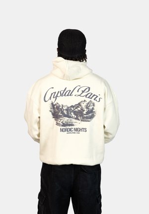 Felpa color crema con cappuccio, caratterizzata da una grafica di montagne e una baita sul retro, con la scritta "Crystal Paris Nordic Nights" sottostante. Accenti neri visibili.