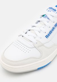 Vit sneaker med mockadetaljer och perforerat tåparti. Har Reebok-logotyp i blått på sidan, runda snören och en strukturerad sula.