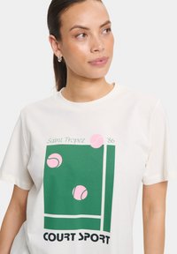 T-shirt crème avec un graphique vert et des balles de tennis roses, avec du texte indiquant "Saint Tropez '86" et "COURT SPORT" en gras noir.