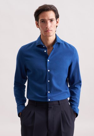 Camicia blu con bottoni in un tessuto liscio e colletto rigido, con bottoni bianchi e maniche lunghe, abbinata a pantaloni scuri.