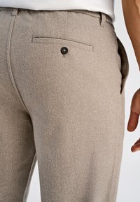 Grijze broek met textuur, een achterzak en een zwarte knoopdetails; de stof heeft een zachte, geweven uitstraling. Getailleerde pasvorm met zijzakken.