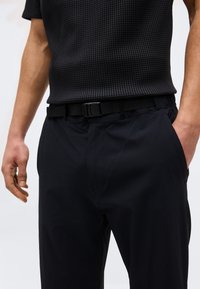 Άνδρας που φοράει μαύρο κοντομάνικο πουκάμισο με υφή, κρυμμένο μέσα σε μαύρο παντελόνι με ρυθμιζόμενο μαύρο υφασμάτινο ζώνη, με το ένα χέρι στην τσέπη.