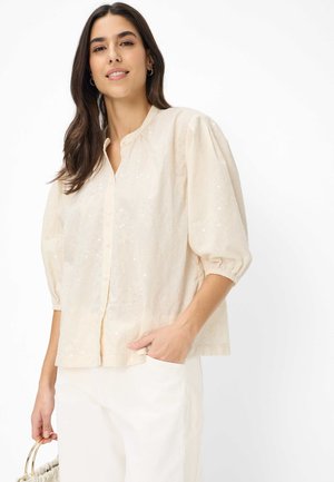 Frau mit langen dunklen Haaren trägt eine cremefarbene, strukturierte Bluse mit Puffärmeln und weiße Hosen, hält eine beige Handtasche mit goldenen Griffen.