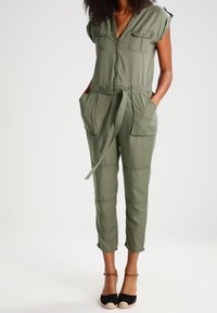 Kobieta ubrana w oliwkowozielony kombinezon z paskiem, krótkimi rękawami i przednimi kieszeniami, w połączeniu z czarnymi espadrylami na obcasie.