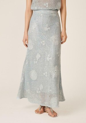 Mujer con falda maxi de crochet azul claro con motivos florales, combinada con sandalias de dedo con joyas sobre un fondo neutro.