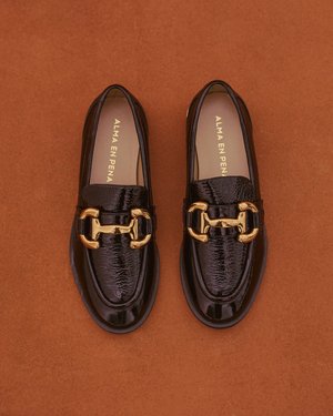 Mocasines de charol negro con un acento de cadena dorada, textura suave, punta redonda y un diseño clásico sin cordones.