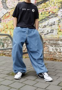 Camisa de algodón negra con detalles de logo, pantalones de mezclilla azul holgados y zapatos deportivos en blanco y negro, contra una pared de graffiti.