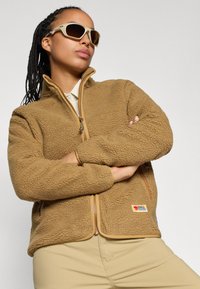 Giacca in pile marrone con superficie texturizzata, chiusura con zip e logo a patch. Occhiali da sole chiari completano l'outfit, indossato in un contesto neutro.