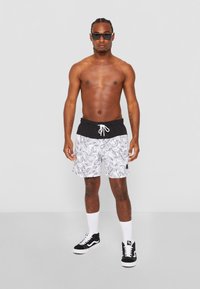 Svart-hvite svømmetights med bladmønster, svart livband, snorstramming, kombinert med svarte sneakers og hvite sokker; modellen er uten skjorte.