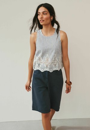 Kobieta o długich, ciemnych włosach, ubrana w bezrękawnik w paski z koronkowymi wykończeniami oraz ciemne krótkie spodenki do kolan, stojąca na tle jednolitego tła.