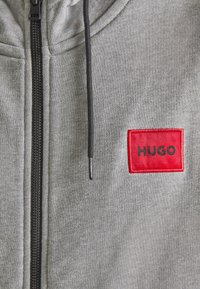 Sudadera gris con cremallera negra, con un parche bordado rojo que lleva el texto "HUGO". La textura de la tela parece suave y de punto.