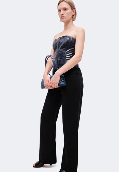 Une femme porte un haut noir bustier texturé, un pantalon à jambes larges et un sac à main bleu devant un fond clair et uni.