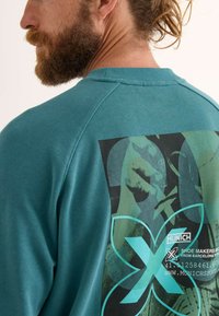 Sudadera de algodón en color verde azulado con un gran estampado gráfico en la parte posterior que presenta formas abstractas y detalles de la marca en acentos negros y turquesa.