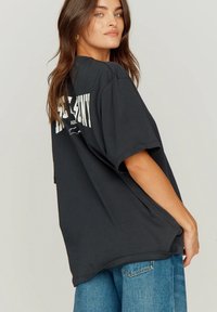 Νεαρή γυναίκα με μακριά καστανά μαλλιά κοιτάζει πάνω από τον ώμο της, φορώντας μια φαρδιά μαύρη μπλούζα με στάμπα και μπλε τζιν, πάνω σε απλό φόντο.