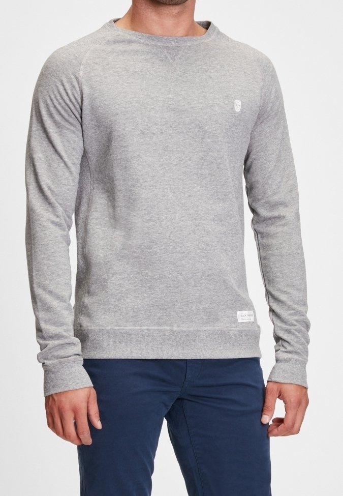 Homme portant un sweat-shirt gris clair ajusté et un pantalon bleu foncé, debout devant un fond blanc uni.