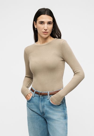 Γυναίκα με μακριά σκούρα μαλλιά φοράει μπεζ ριμπ πλεκτό μακρυμάνικο τοπ, μπαίνει μέσα σε μπλε τζιν ψηλόμεσα με καφέ ζώνη, χέρια στις τσέπες.