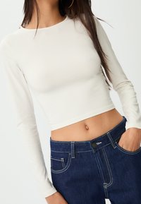 Dugi rukav, skraćeni top u off-white tkanini s okruglim izrezom. Uparen s tamnoplavim trapericama, s vidljivim šavovima i detaljima džepova.