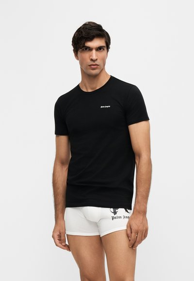 Άνδρας με σκούρα μαλλιά φοράει μαύρο T-shirt Palm Angels και λευκά boxer Palm Angels, στέκεται μπροστά σε απλό φωτεινό φόντο.