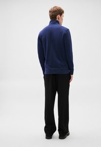 Tumši zils džemperis ar augstu apkakli, izgatavots no mīksta materiāla. Komplektā ir garas piedurknes un brīvs siluets, kombinēts ar melnām biksēm un apaviem.